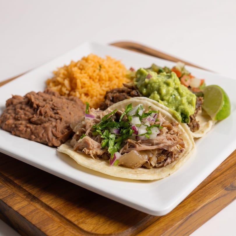 A plate of carnitas tacos, refried beans, Mexican rice, guacamole, and a lime wedge.